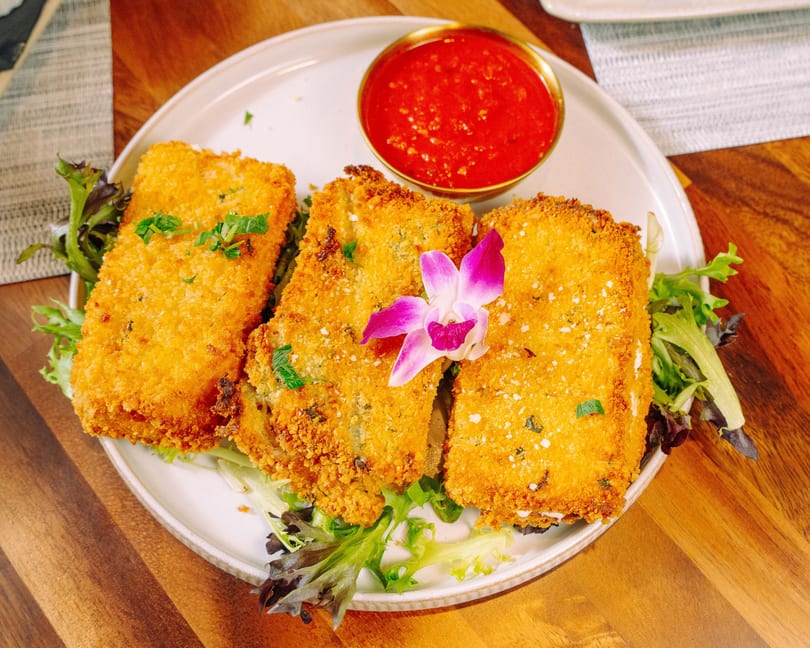 Three golden-brown fried ravioli on a bed of greens with a side of red marinara sauce.