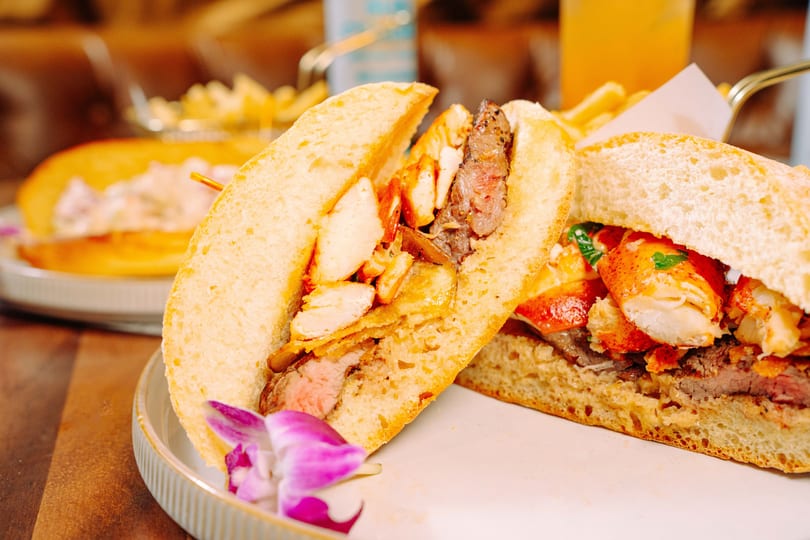 Two gourmet sandwiches, one with steak and chicken, the other with lobster and steak, on a white plate.