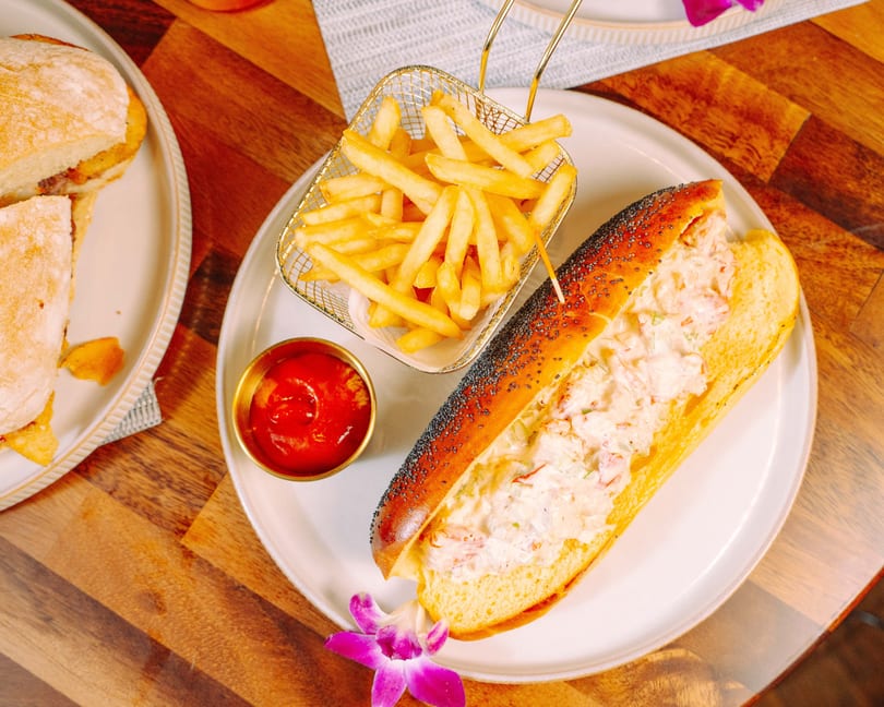 Lobster roll with fries and ketchup on a white plate, garnished with a purple orchid.