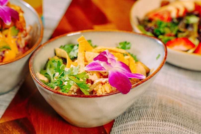 A colorful bowl of rice, cilantro, tortilla strips, and a purple orchid on a wooden table.