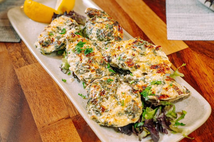 Baked oysters topped with cheese, spinach, and herbs, garnished with lemon wedges on a white platter.