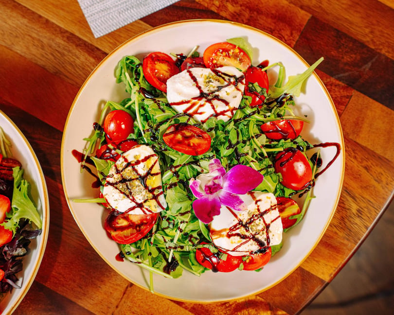 Caprese salad with fresh mozzarella, cherry tomatoes, arugula, balsamic glaze, and an orchid garnish.